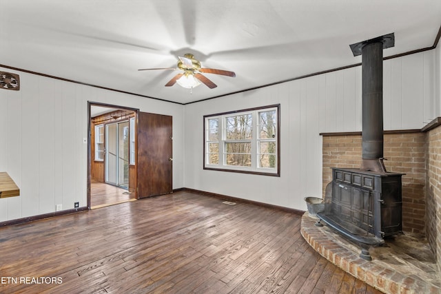 unfurnished living room with baseboards, wood-type flooring, ceiling fan, ornamental molding, and a wood stove