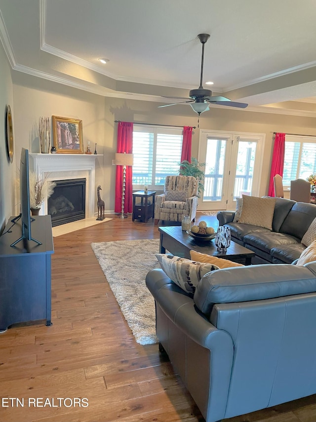 living area featuring light wood finished floors, a raised ceiling, a ceiling fan, a fireplace with flush hearth, and crown molding