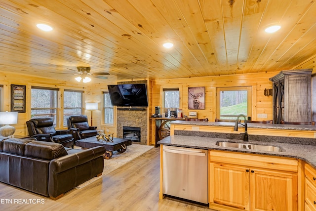 kitchen with light wood-type flooring, a stone fireplace, wood walls, ceiling fan, and stainless steel dishwasher