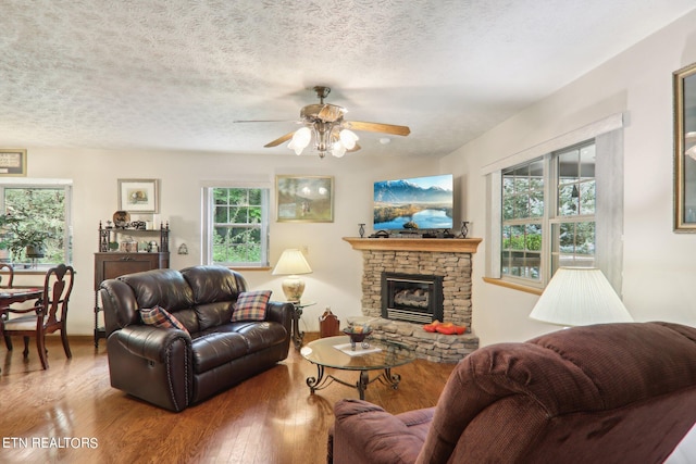 living room with a textured ceiling, ceiling fan, a stone fireplace, and wood-type flooring