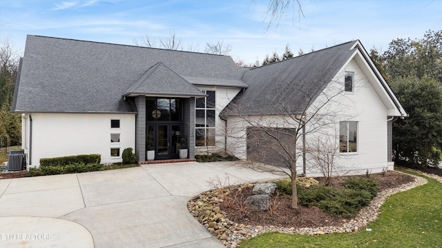 french country inspired facade with brick siding, roof with shingles, an attached garage, cooling unit, and driveway