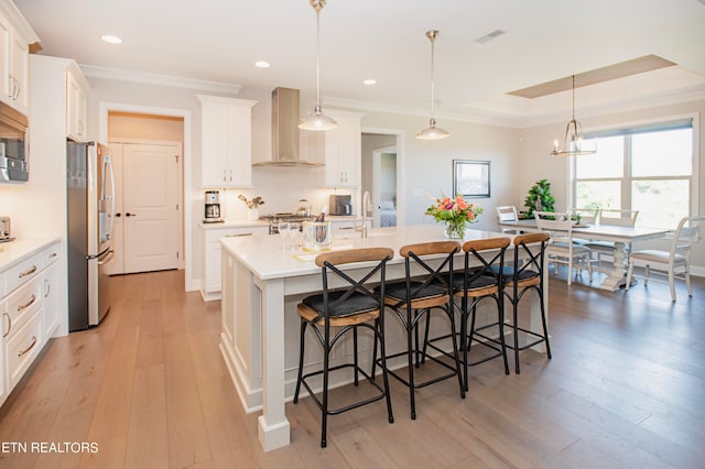 kitchen featuring light hardwood / wood-style flooring, appliances with stainless steel finishes, wall chimney range hood, pendant lighting, and a kitchen island with sink