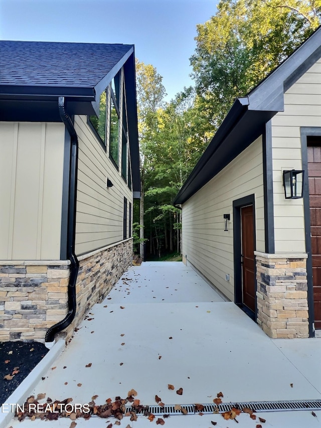 view of property exterior featuring a patio, stone siding, board and batten siding, and roof with shingles