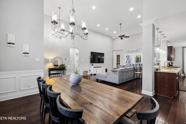 dining area with ceiling fan with notable chandelier, dark wood finished floors, a wainscoted wall, a towering ceiling, and a decorative wall