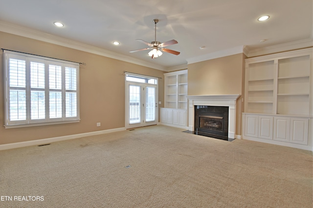 unfurnished living room with a tiled fireplace, crown molding, built in shelves, light colored carpet, and ceiling fan