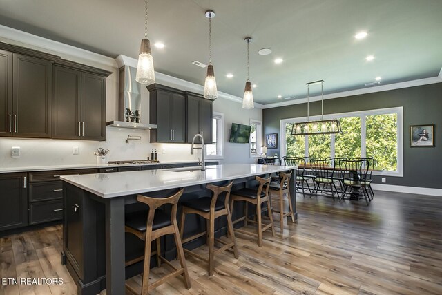 kitchen featuring dark hardwood / wood-style flooring, stainless steel gas cooktop, a kitchen island with sink, sink, and wall chimney range hood