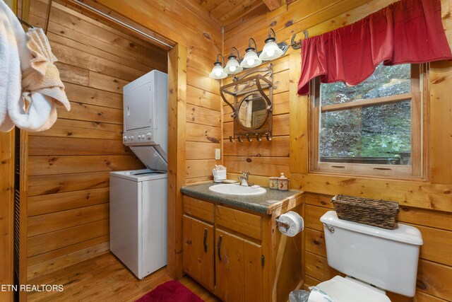 bathroom featuring stacked washer / dryer, wooden walls, vanity, and toilet