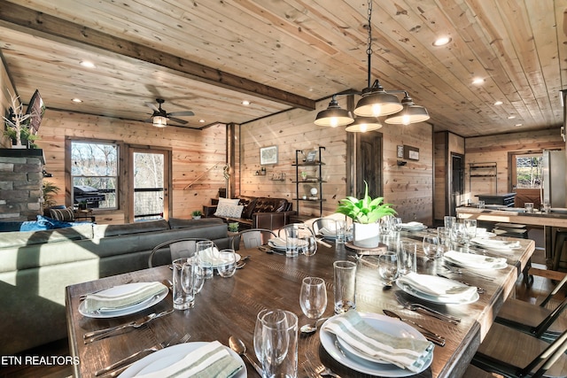 dining area featuring wooden walls, ceiling fan, a stone fireplace, wood ceiling, and beam ceiling