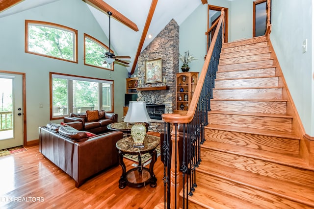 living room with ceiling fan, beamed ceiling, a stone fireplace, and light hardwood / wood-style floors