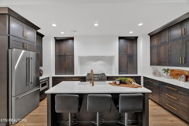 kitchen with sink, an island with sink, dark brown cabinets, appliances with stainless steel finishes, and light wood-type flooring