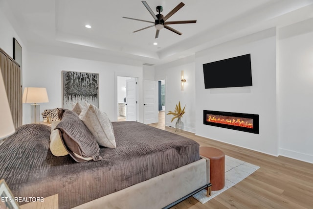 bedroom featuring light wood-type flooring, a tray ceiling, ceiling fan, and ensuite bathroom