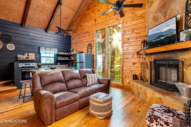 living room with beamed ceiling, a stone fireplace, wooden walls, high vaulted ceiling, and light wood-type flooring