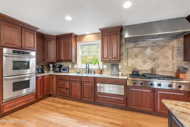 kitchen with light hardwood / wood-style floors, light stone counters, wall chimney exhaust hood, stainless steel appliances, and sink