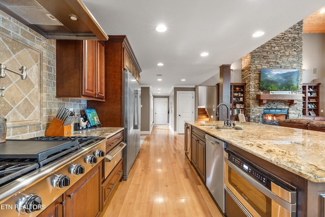 kitchen featuring sink, light hardwood / wood-style flooring, stainless steel appliances, exhaust hood, and a stone fireplace