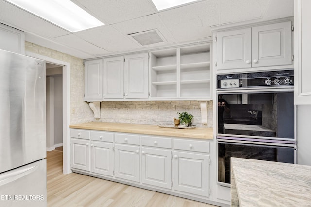 kitchen featuring white cabinets, stainless steel refrigerator, black double oven, light wood-type flooring, and decorative backsplash