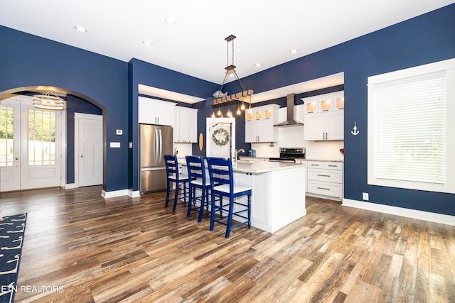 kitchen with a center island with sink, stainless steel fridge, wall chimney range hood, decorative light fixtures, and white cabinets