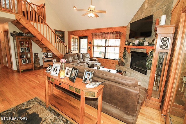 living room with ceiling fan, wood walls, high vaulted ceiling, a fireplace, and light hardwood / wood-style floors