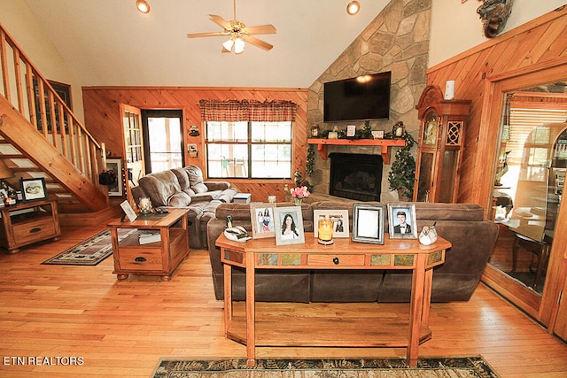 living room with high vaulted ceiling, a stone fireplace, light hardwood / wood-style flooring, ceiling fan, and wooden walls