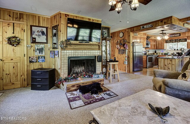 living room featuring a brick fireplace, wood walls, a textured ceiling, ceiling fan, and light colored carpet