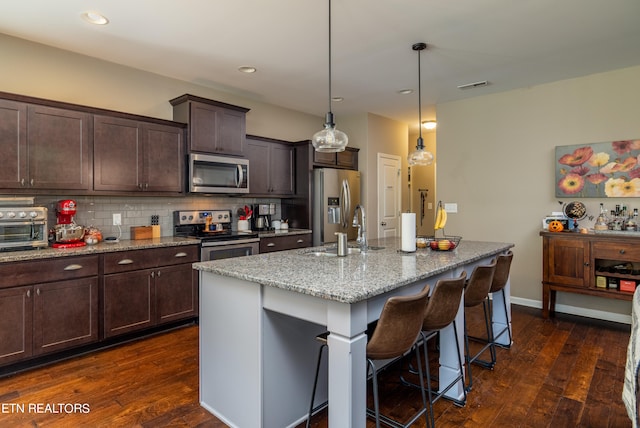 kitchen with appliances with stainless steel finishes, an island with sink, light stone countertops, dark hardwood / wood-style floors, and sink