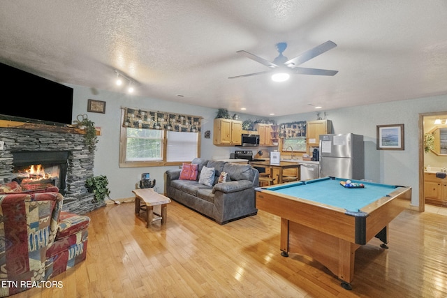 recreation room featuring a textured ceiling, light hardwood / wood-style floors, billiards, a stone fireplace, and ceiling fan