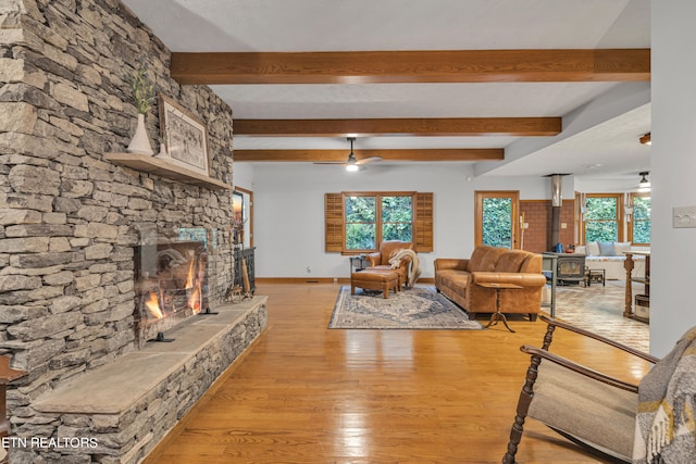 living room with light hardwood / wood-style flooring, ceiling fan, a stone fireplace, beamed ceiling, and a wood stove