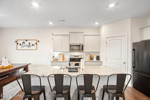 kitchen with dark hardwood / wood-style floors, a kitchen bar, stainless steel appliances, a center island with sink, and light stone countertops