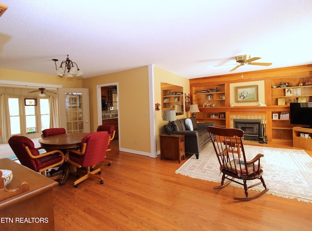 dining area featuring ceiling fan with notable chandelier, baseboards, a textured ceiling, light wood-style floors, and a tiled fireplace