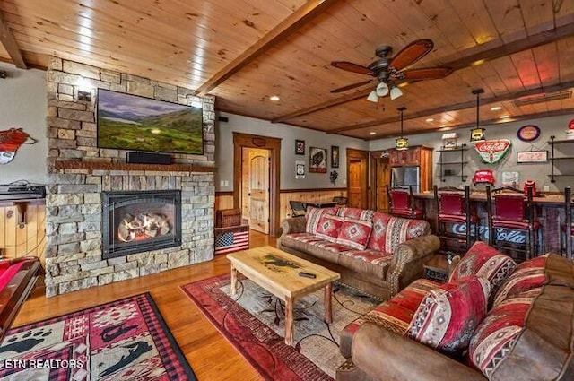 living room featuring ceiling fan, beam ceiling, hardwood / wood-style floors, a stone fireplace, and wood walls