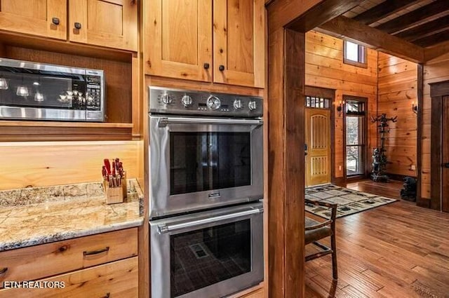 kitchen featuring wood walls, light wood-type flooring, light stone countertops, appliances with stainless steel finishes, and beamed ceiling