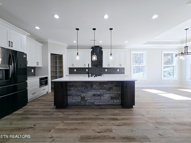 kitchen featuring a kitchen island with sink, white cabinetry, wall chimney exhaust hood, light countertops, and black refrigerator with ice dispenser