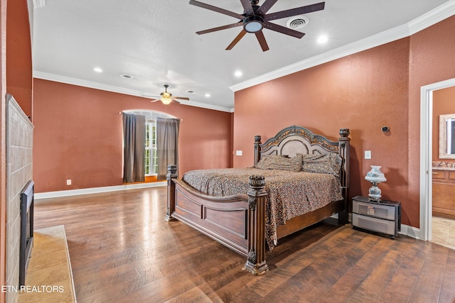 bedroom with hardwood / wood-style flooring, a tiled fireplace, connected bathroom, crown molding, and ceiling fan