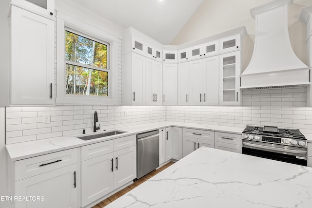 kitchen with light stone countertops, lofted ceiling, custom range hood, appliances with stainless steel finishes, and a sink