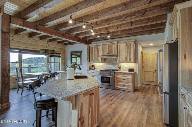 kitchen featuring light stone counters, beamed ceiling, a kitchen island with sink, dark wood-type flooring, and appliances with stainless steel finishes