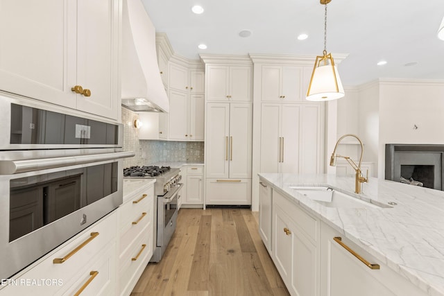 kitchen featuring premium range hood, stainless steel appliances, sink, decorative light fixtures, and light hardwood / wood-style floors