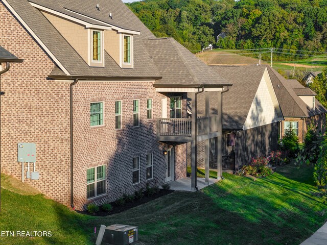 back of house featuring a shingled roof, a balcony, a yard, and brick siding