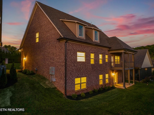 back house at dusk featuring a patio area, a balcony, and a lawn