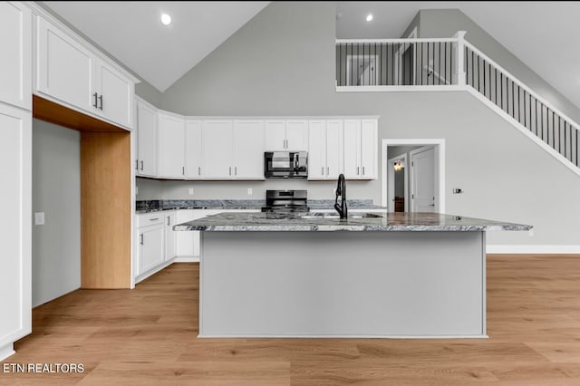 kitchen featuring white cabinets, a center island with sink, high vaulted ceiling, stainless steel appliances, and dark stone counters