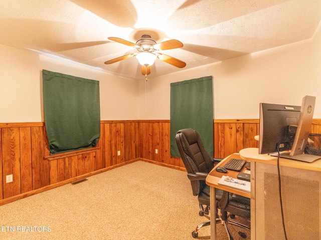 carpeted home office with ceiling fan, wood walls, and a textured ceiling