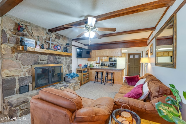 living room with a stone fireplace, beam ceiling, light colored carpet, a textured ceiling, and ceiling fan