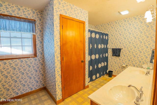 full bath with a sink, double vanity, a textured ceiling, and wallpapered walls