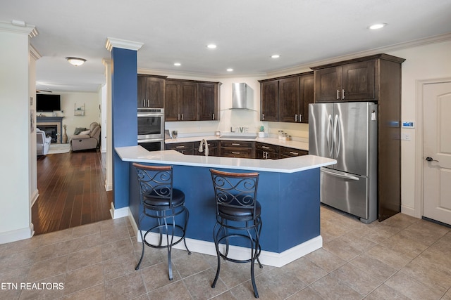 kitchen with wall chimney range hood, appliances with stainless steel finishes, light wood-type flooring, a kitchen bar, and crown molding