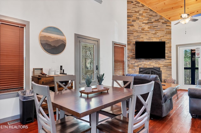 dining area featuring wooden ceiling, dark wood-type flooring, high vaulted ceiling, a stone fireplace, and ceiling fan