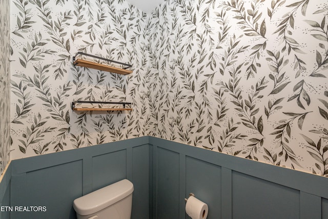 half bath featuring toilet, wallpapered walls, a decorative wall, and wainscoting