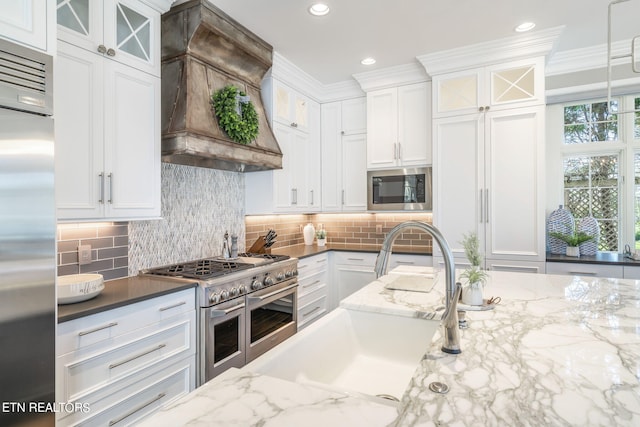 kitchen with decorative backsplash, built in appliances, custom range hood, hanging light fixtures, and white cabinets