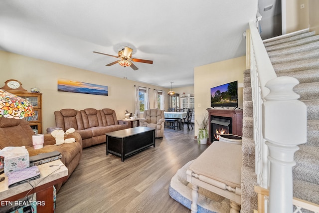 living room with a ceiling fan, a lit fireplace, stairway, and wood finished floors