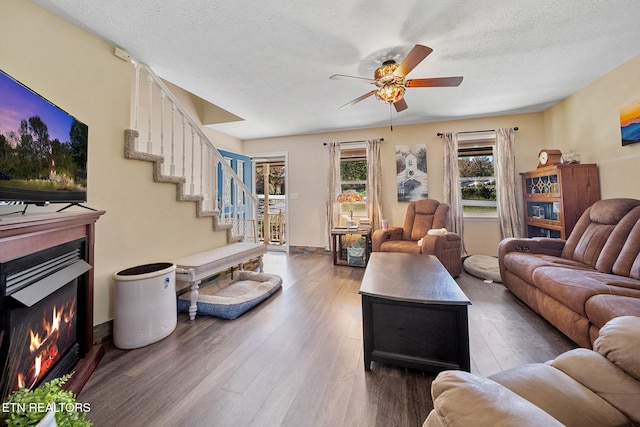 living room with a warm lit fireplace, a ceiling fan, dark wood-style flooring, stairs, and a textured ceiling