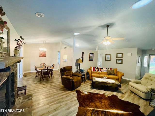 living room featuring ceiling fan, vaulted ceiling, light wood-type flooring, a stone fireplace, and a textured ceiling