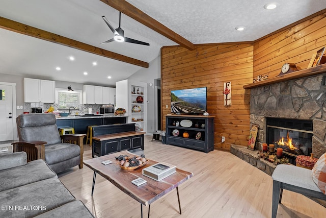 living room with ceiling fan, vaulted ceiling with beams, a fireplace, light hardwood / wood-style floors, and wooden walls