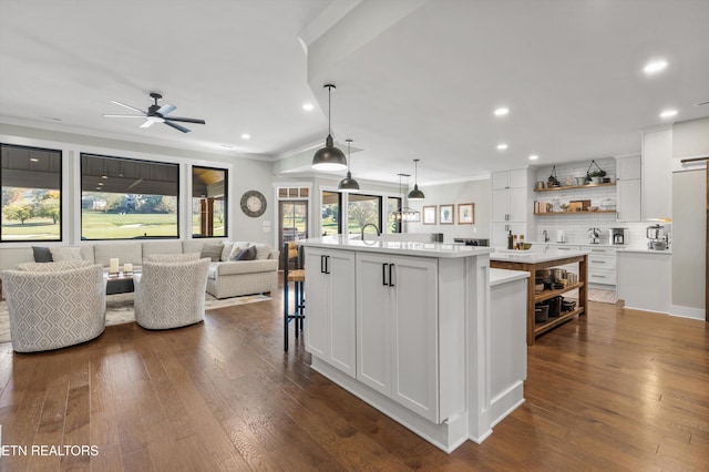 kitchen featuring backsplash, a kitchen island with sink, crown molding, decorative light fixtures, and white cabinetry